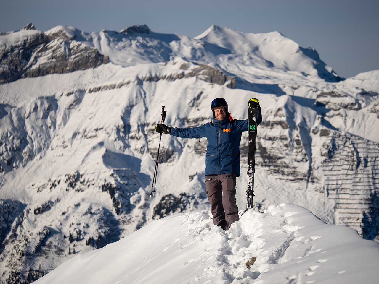 Skifahrer steht auf Bergspitze, hält seine Skiern in Händen und lächelt in die Kamera